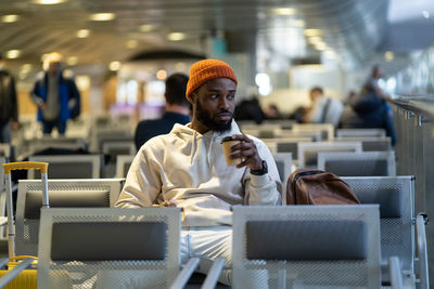 Young african man drinking coffee and eating sandwich while waiting for flying at airport terminal