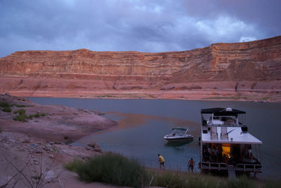 Ferry moored in lake powell by rocky mountains against cloudy sky