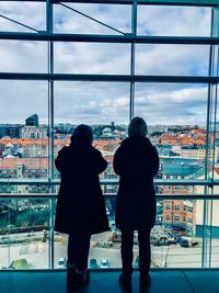 Rear view of people standing by a window enjoying the city view