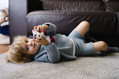 Cute little boy lying on carpet and playing with toy car in living room at home