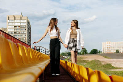 Two teenage girls walk together through the stands of the school stadium, talking, holding hands