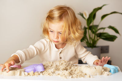 Cute happy caucasian,blonde,curly-haired toddler,baby girl playing with kinetic sand indoors