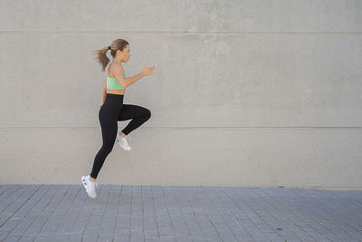 Side view of athletic woman in activewear jumping above ground in street while warming up body during workout