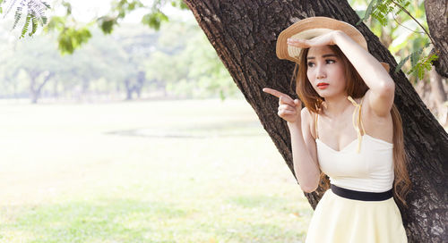 Portrait of beautiful young woman wearing hat while standing by tree trunk