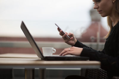 Side view of woman using mobile phone and laptop while sitting at sidewalk cafe