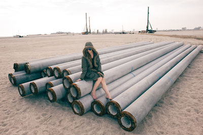 Woman sitting on metallic pipes at beach against sky