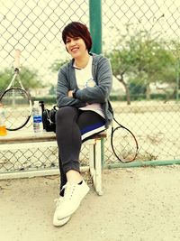 Young woman sitting on bench by chainlink fence