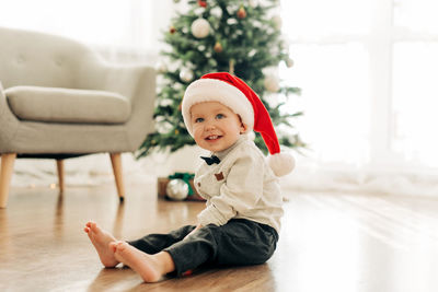 A laughing boy in a santa claus hat sits on the floor against the background of a christmas tree