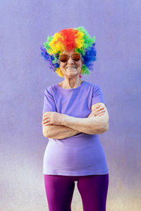 Content senior female athlete in modern sunglasses and bright wig standing with folded arms on purple background
