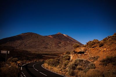 Road leading towards mountains against clear blue sky