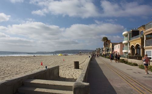 Rear view of people walking on street by beach against cloudy sky