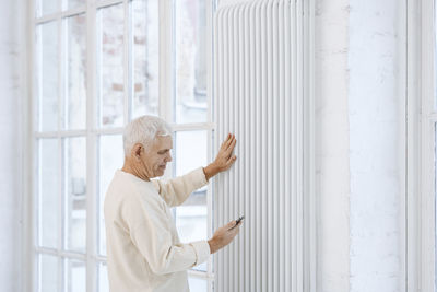 Senior man using smart phone touching radiator at home