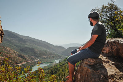 Man sitting on rock looking at mountains against sky