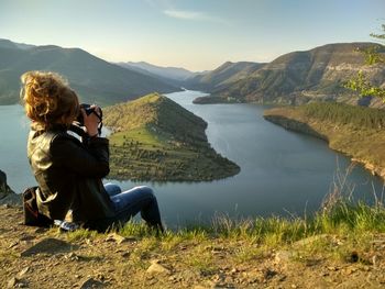 Rear view of man photographing at lake against mountains