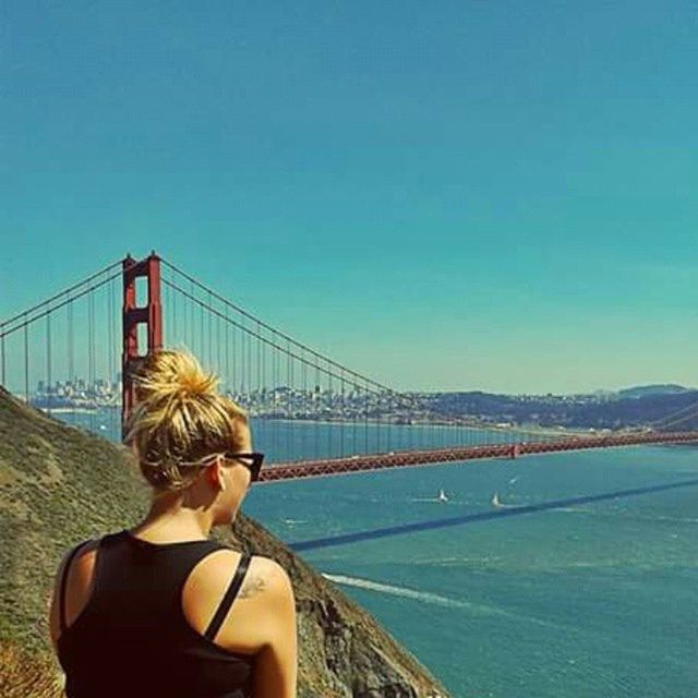 clear sky, lifestyles, leisure activity, water, young adult, connection, sea, copy space, rear view, young women, person, bridge - man made structure, blue, suspension bridge, sitting, standing, built structure, casual clothing