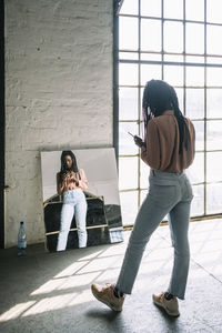 Full length rear view of female it professional using smart phone with reflection in mirror at creative office