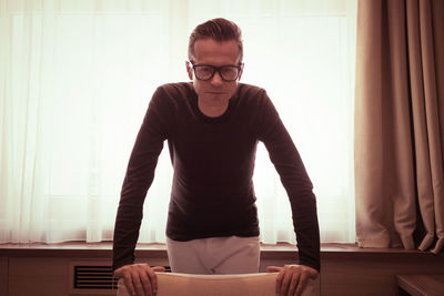 Pensive mid adult man thinking while standing next to window and leaning on armchair.