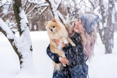 Young woman carrying dog while crouching in snow during winter