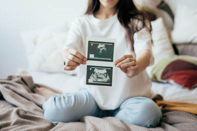 A pregnant woman, sitting on a bed in home clothes, holds an ultrasound scan of a human fetus.