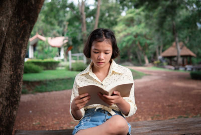 Young woman sitting on book against tree trunk