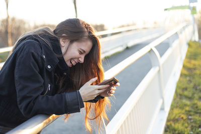 Young woman using mobile phone while sitting on railing