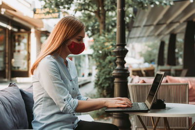 Side view of woman wearing mask using laptop at cafe