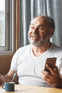 Handsome mature man laughs while has video call with cellphone and headphones at home.