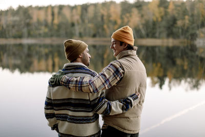 Male friends talking to each other while standing with arm around against lake