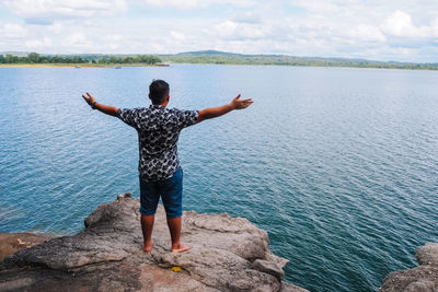 Rear view of man with arms outstretched standing on rock by sea against sky