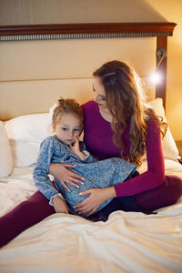 Mother in purple clothes with a sad dissatisfied daughter in a dress is sitting on a bed in a hotel