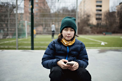 Portrait of boy with mobile phone wearing warm clothing while sitting against soccer field in city