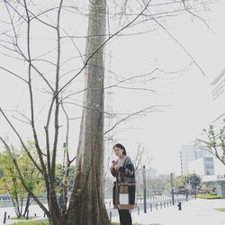 Portrait of young woman standing on bare tree against sky
