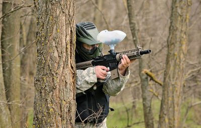 Teenage boy playing paintball against bare trees in forest