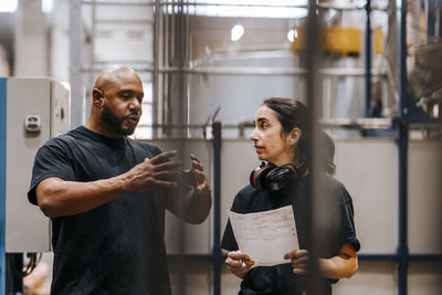 Mature male worker gesturing while discussing strategy with female coworker wearing headphones standing in manufacturing