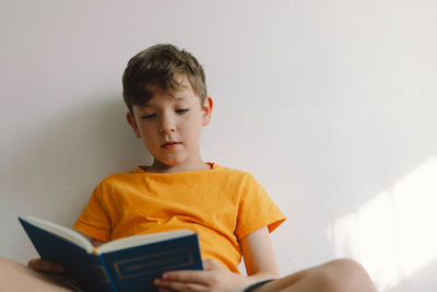 A cute boy wearing an orange t-shirt is sitting on a soft ottoman reading a book