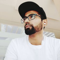 Low angle view of bearded young man wearing eyeglasses and cap listening music on headphones