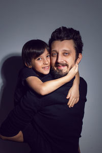 Boy son  sitting on father with a beard and hair back in studio on gray background in black clothes