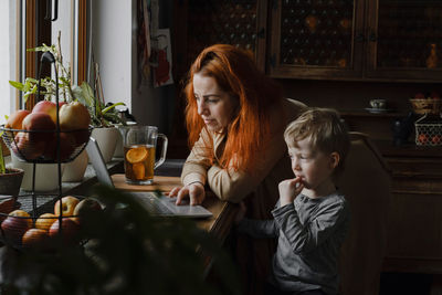 Ginger haired woman on kitchen with child looking in laptop. vintage house, cozy family life. boy