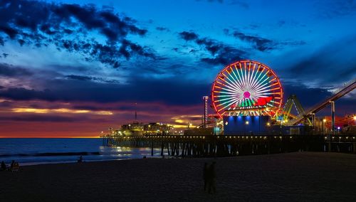 Illuminated ferris wheel at beach against sky during sunset