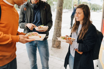 Smiling young woman enjoying meal with male friends while standing on street by food truck