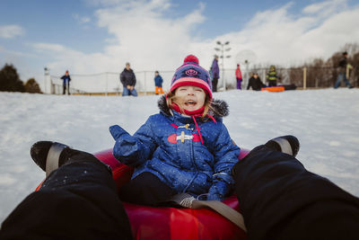 Low section of mother with daughter tobogganing on snow covered field against cloudy sky