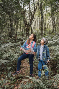 Ethnic girl with paper guide looking away against brother with binoculars among fern plants in summer woods