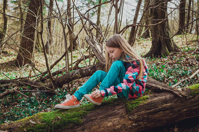 Side view of little girl tying shoelace while sitting on log in forest