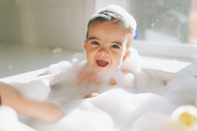 Cute happy baby boy taking bubble bath in kitchen sink at home