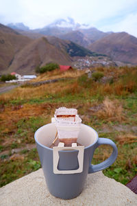 Cup of portable drip coffee being prepared at outdoor terrace with blurry village view in background