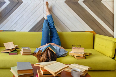 Young woman reading on the sofa at the living room with a lot of closed and opened books around. 