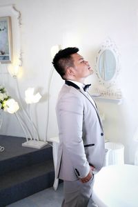 Side view of bridegroom wearing gray suit standing with eyes closed against wall