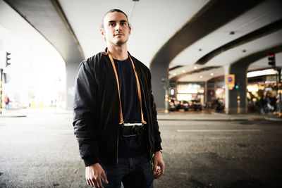 Young man with camera standing on street below overpass in city