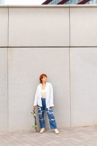 Full body of female in casual wear with skateboard looking away while standing on gray background in city