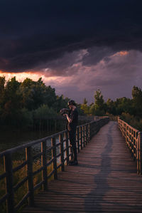 Man using camera while standing on footbridge against sky